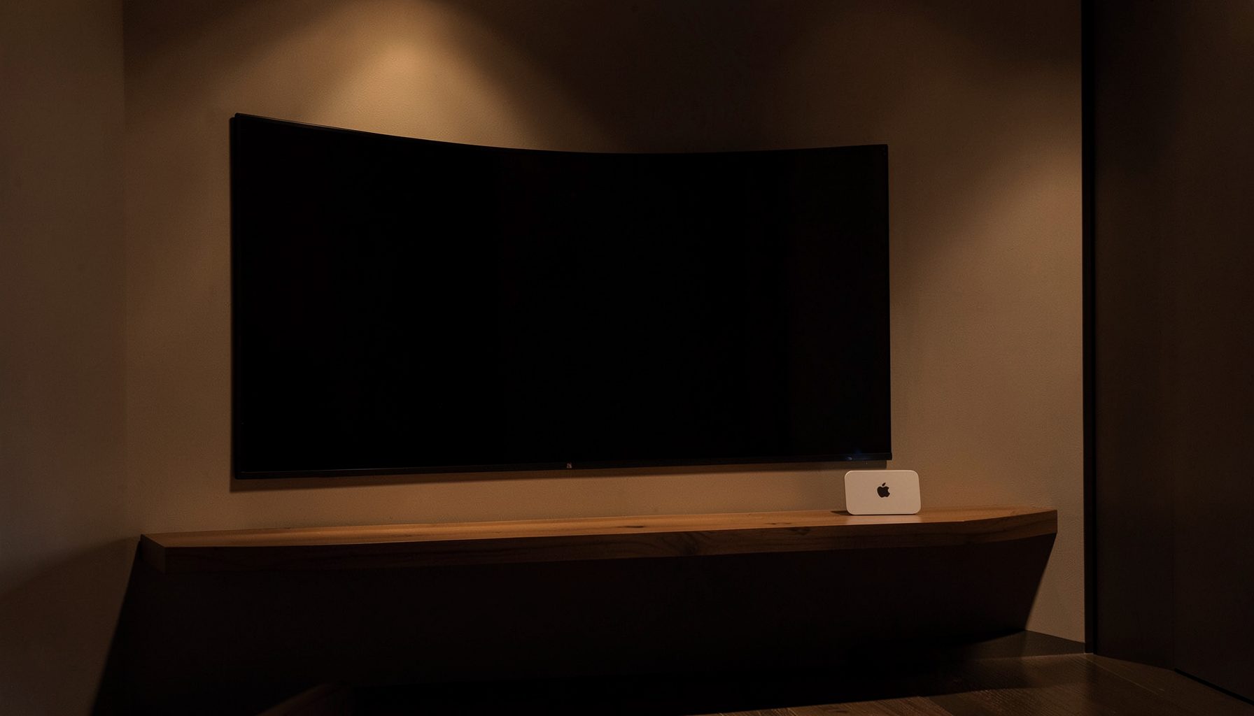A minimalist home theater setup with a wall-mounted TV and a small Apple TV device on a floating shelf, warm ambient lighting and deep shadow tones