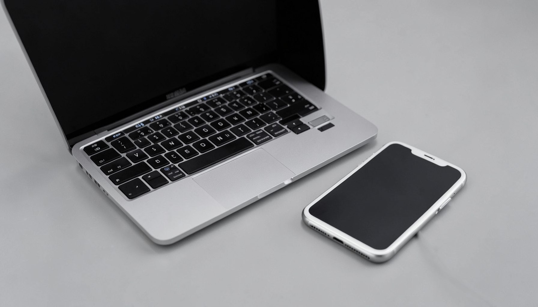 A modern laptop next to an iPhone connected via USB cable, neutral gray and silver tones